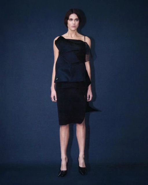 A female fashion model poses in a black asymmetrical top and pencil skirt against a blue background in a full shot in a studio.