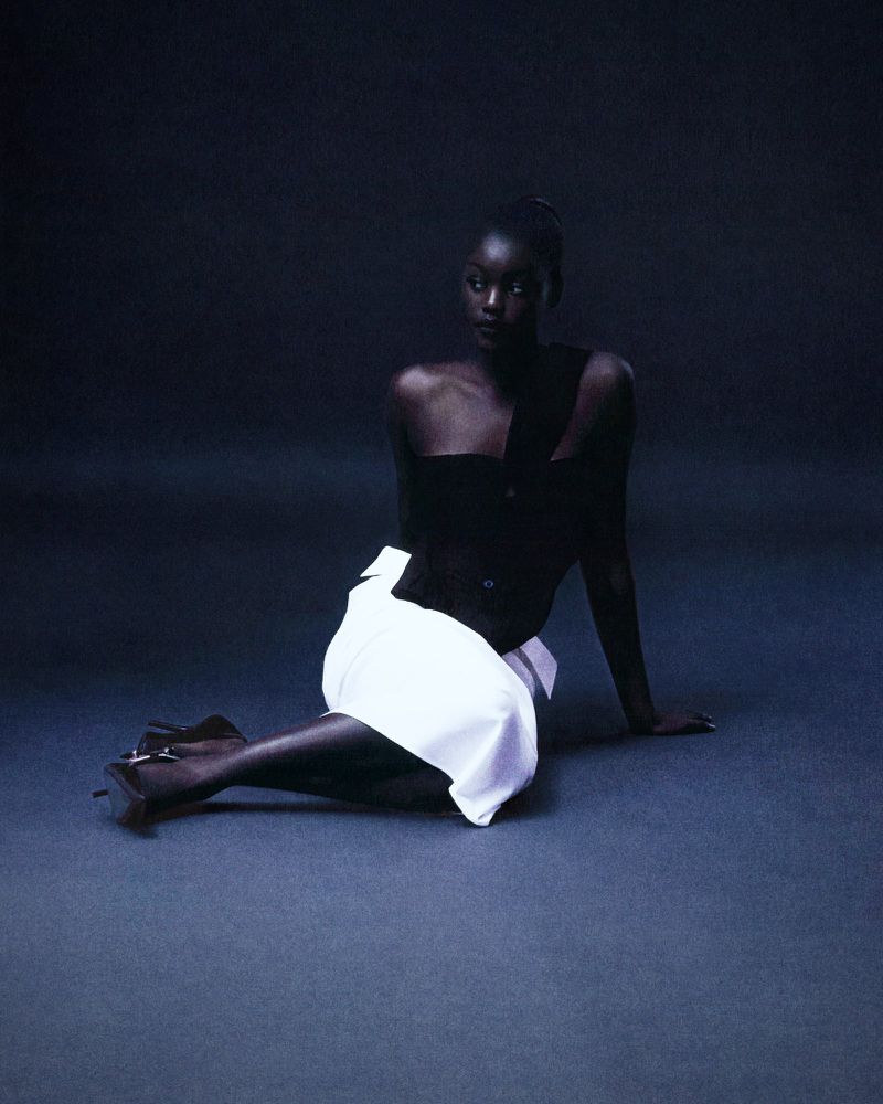 Elegant fashion shot of a Black woman posing in a dark studio, wearing a black top and white skirt. She is looking off to the side.