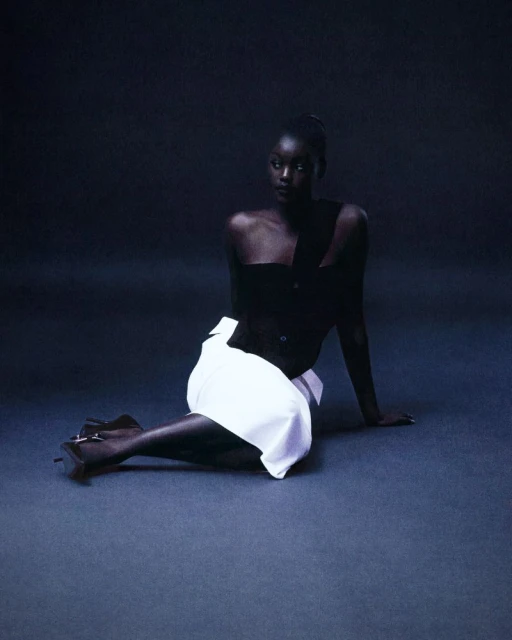 Elegant fashion shot of a Black woman posing in a dark studio, wearing a black top and white skirt. She is looking off to the side.
