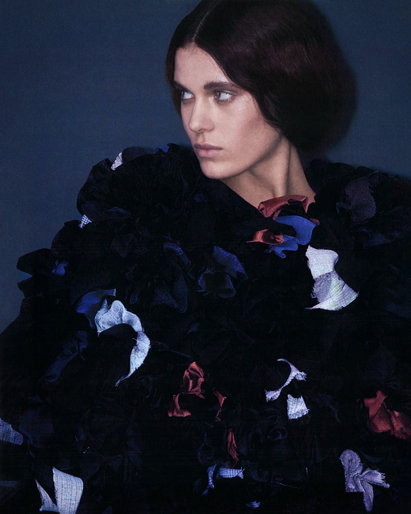 Portrait of a woman with dark hair looking away, wearing a black ruffled garment against a dark blue background in a studio.
