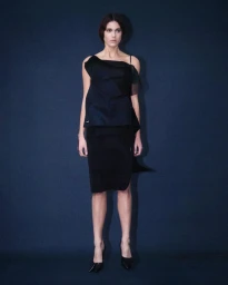 A female model wearing a dark structured top and matching skirt standing against a dark blue textured background