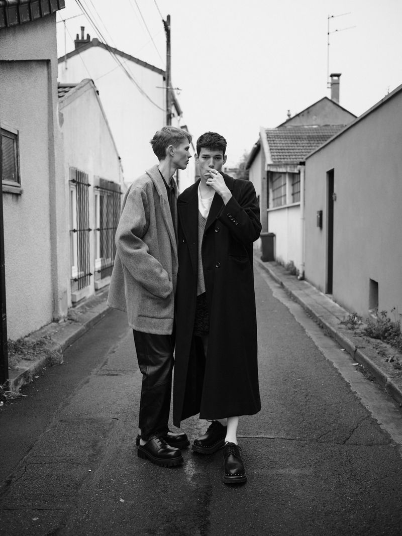 Two young men stand close on a wet street in a black and white editorial fashion shot featuring overcoats and a narrow depth of field.
