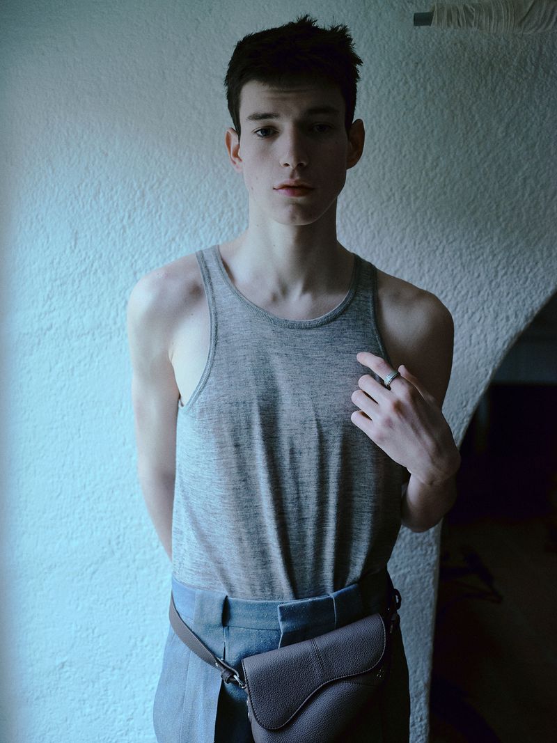 Portrait of an adult male wearing a grey tank top and blue pants, accessorized with a brown leather fanny pack against a white wall.