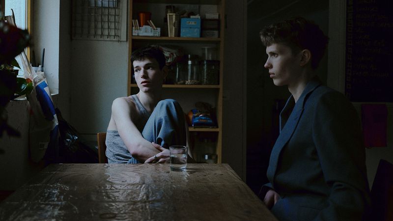 Two young men sit at a table in a dimly lit room. One is wearing a suit jacket, while the other is in a tank top. A glass of water sits on the table.