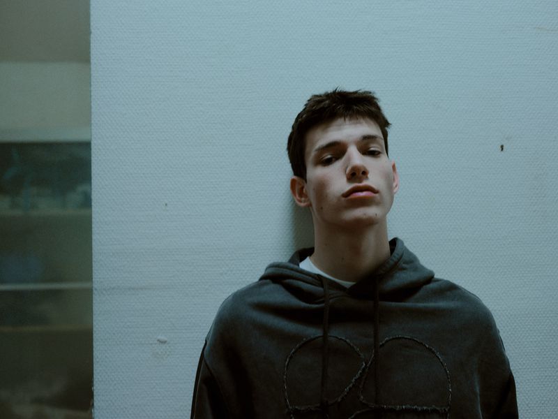 Indoor portrait of a young adult male in a hooded sweatshirt, gazing into the camera against a muted gray wall backdrop.