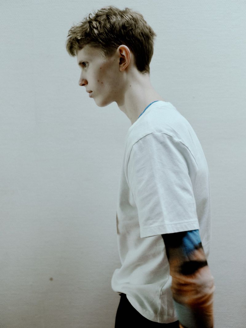 Profile shot of a slender young adult man with short blond hair and a white tee shirt standing against a white wall, with tattooed arm.