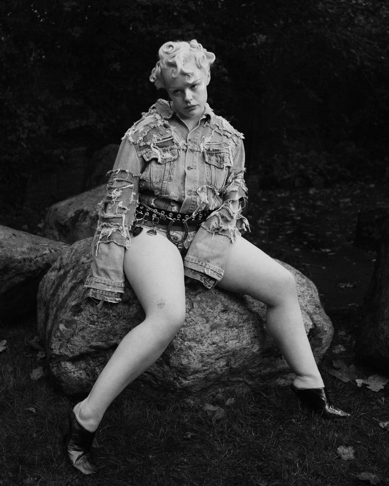 Black and white portrait of an adult female wearing a denim jacket and sitting on a rock, outdoors in a natural environment.