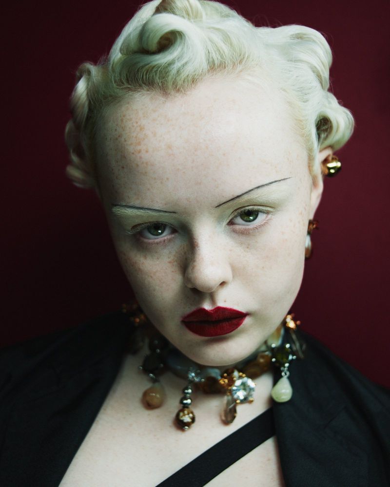 Close-up studio portrait of a young adult female with vintage-style blonde hair, dark red lipstick, and statement gemstone jewelry against a dark red backdrop.