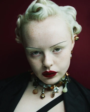 Close-up studio portrait of a young adult female with vintage-style blonde hair, dark red lipstick, and statement gemstone jewelry against a dark red backdrop.