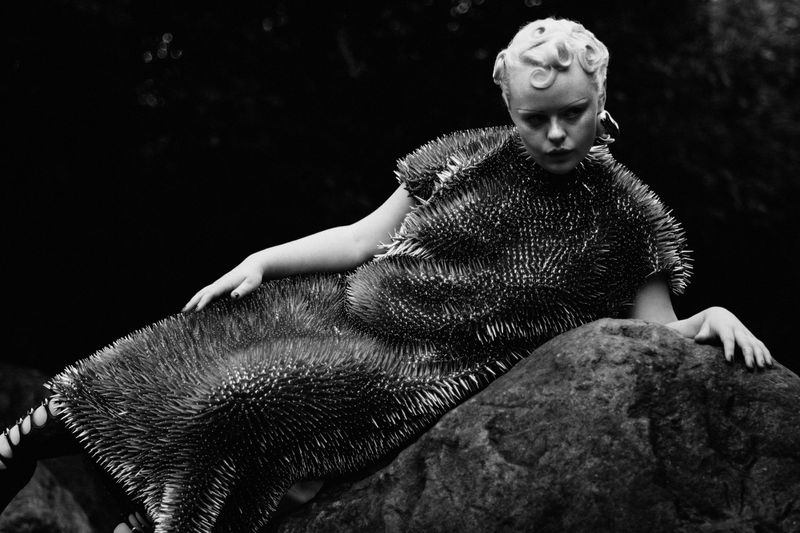 A woman in a spiked dress poses on a rock. The black-and-white image features a light-skinned adult female in a unique garment, indoors.