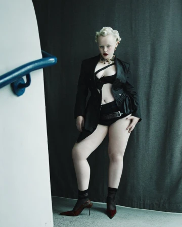 Woman in lingerie, oversized blazer, and heels poses in a dimly lit studio with a white wall and a dark backdrop.