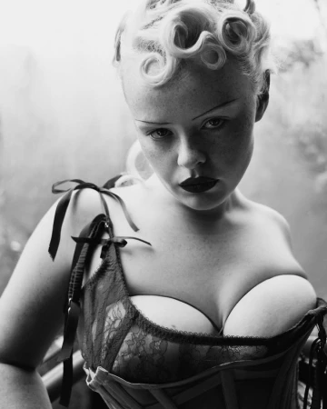Black and white studio portrait of a young adult woman with stylized blonde hair, dark makeup, and a lace bustier corset top.