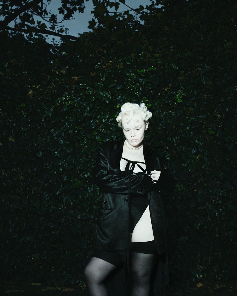 Woman in lingerie and black coat poses outdoors against a dark green foliage backdrop under moody lighting. Fashion editorial image.