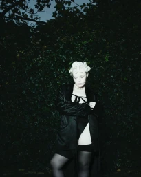 Model with platinum blonde hair wearing an oversized black blazer, black lingerie, and sheer stockings against dark leafy foliage.
