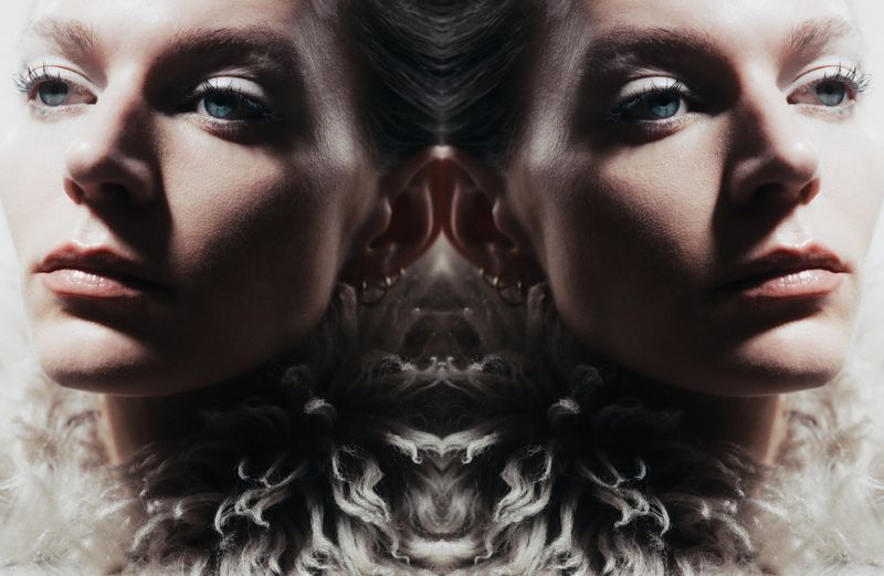 A mirrored portrait showcases a fair-skinned woman with blue eyes, fair hair, and light makeup against a dark background with a furry collar.