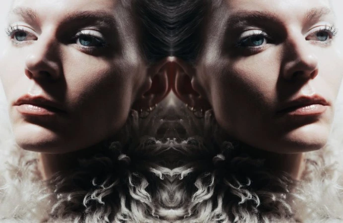 A mirrored portrait showcases a fair-skinned woman with blue eyes, fair hair, and light makeup against a dark background with a furry collar.