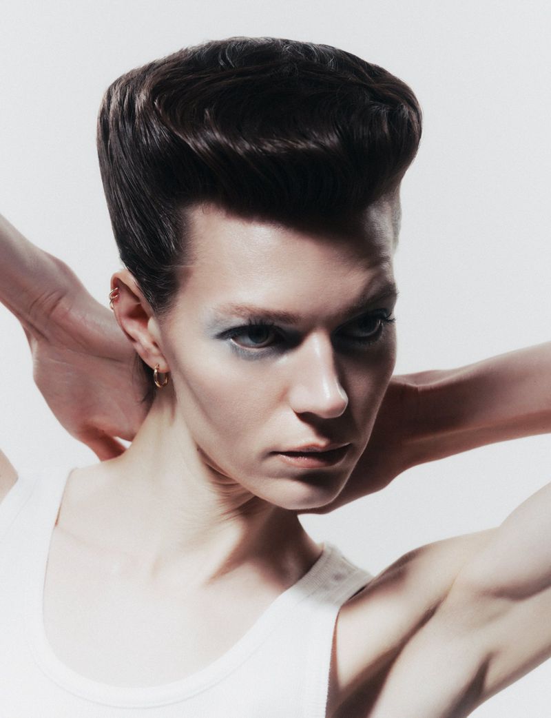 Headshot of androgynous person with stylized dark brown pompadour, hands behind neck, wearing a white tank top against a white background.