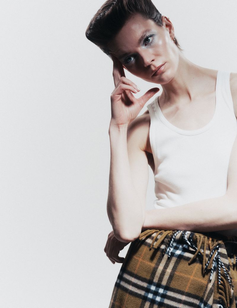 Studio portrait of a young woman with blue eyeshadow, wearing a white tank top and plaid skirt against a white backdrop.
