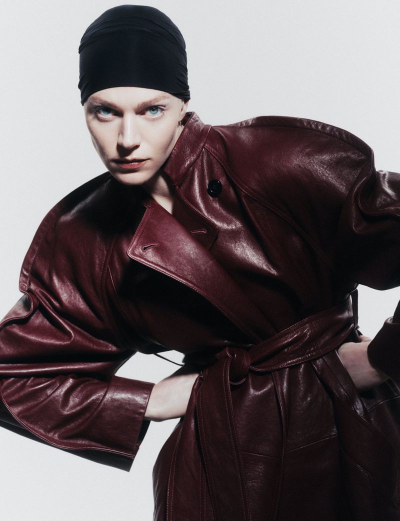 Fashion portrait of a woman with fair skin wearing a black headscarf and a burgundy leather trench coat, posing indoors.