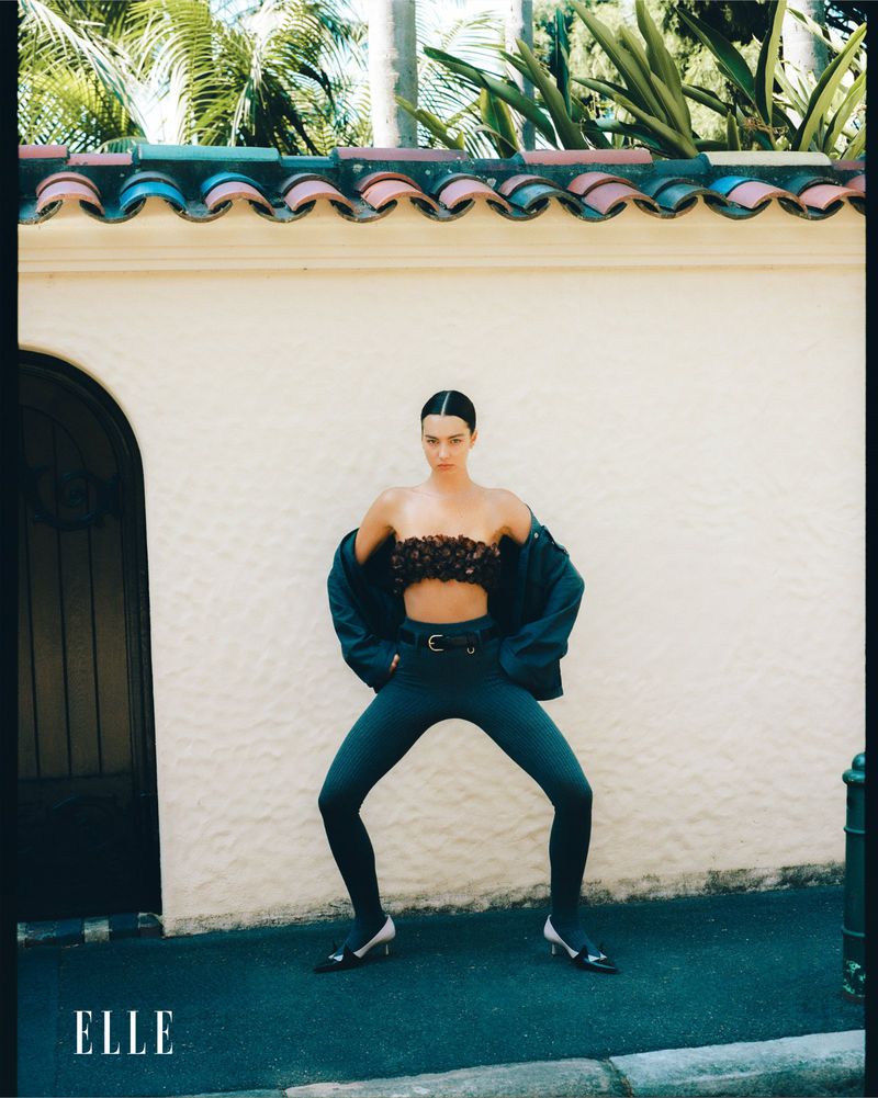 A woman posing in a black bra top and pants near a wall on a sunny day, with palm trees in the background. Fashion editorial.