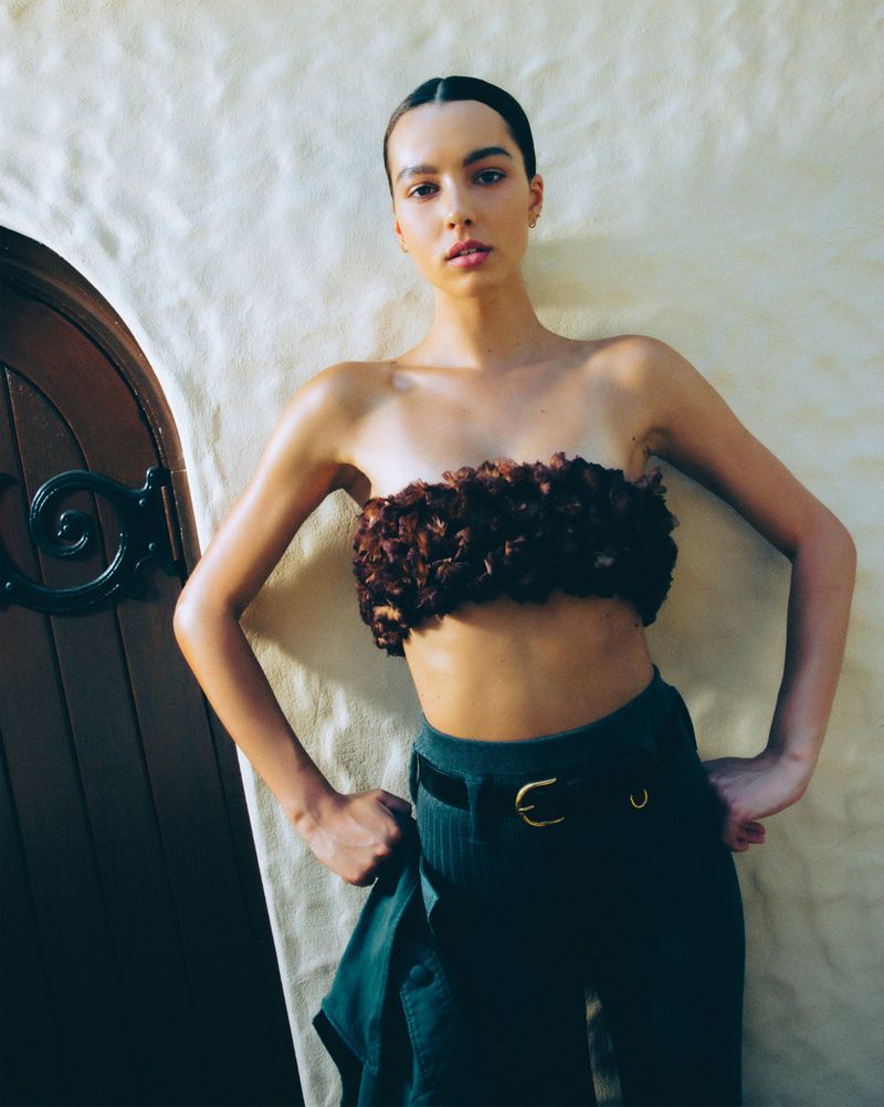 A young adult woman poses indoors in a floral tube top and dark pinstripe pants with a partly opened wooden door in the background.