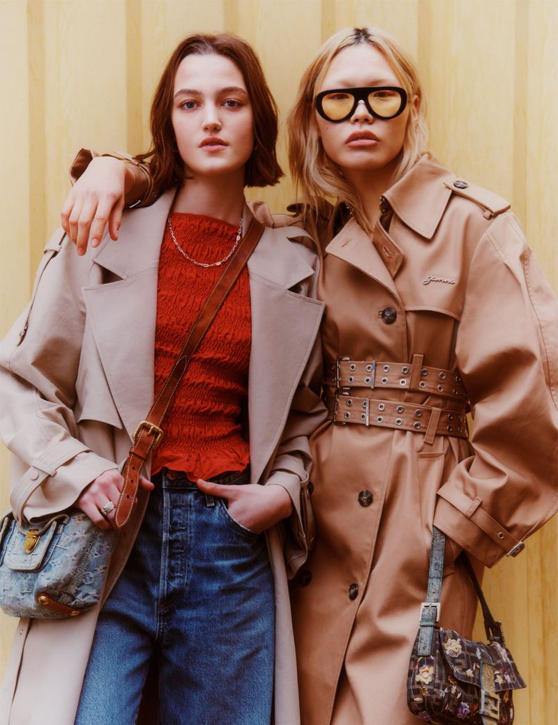 Two women pose in trench coats and jeans against a striped wall, each carrying a handbag in a fashion editorial style photo.
