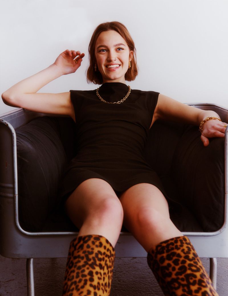 Smiling woman in a black dress sits in a metal-frame chair, wearing leopard-print boots, jewelry, against a white backdrop.