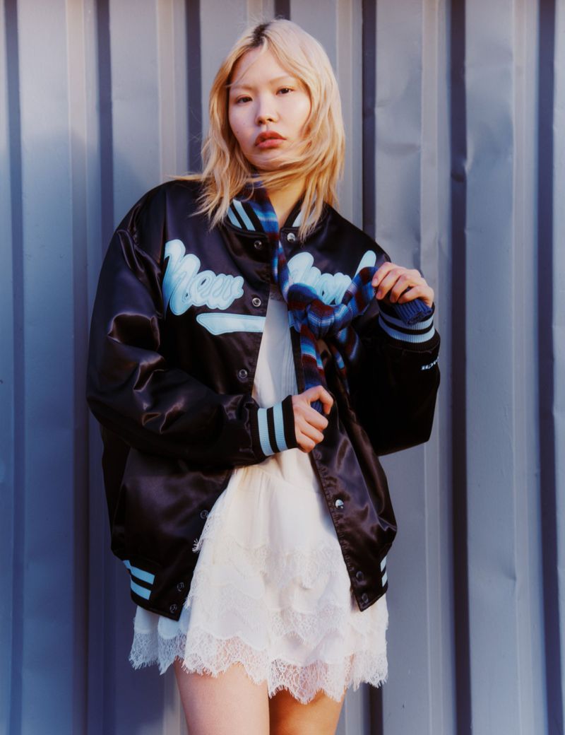 Asian woman with blonde hair wearing a black and blue bomber jacket, standing against a corrugated metal wall. Fashion, style, and portrait.