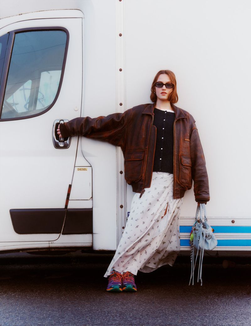 Young woman in stylish brown leather jacket and a floral skirt poses near a white truck on an outdoor asphalt surface.