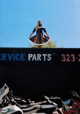 Woman poses in a patterned jacket and skirt, standing atop a dumpster filled with scrap metal, under a clear blue sky outdoors.