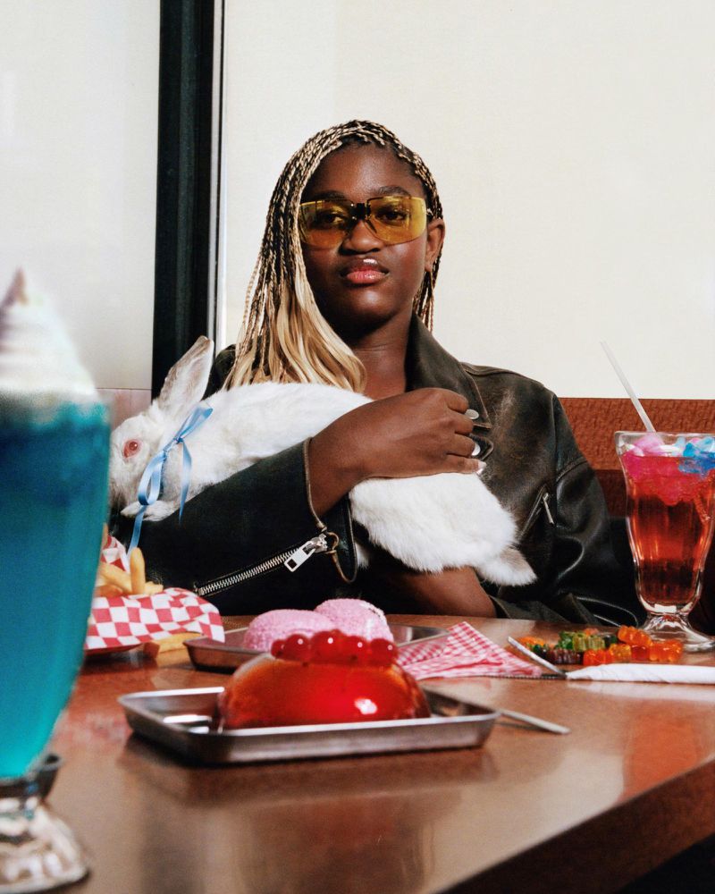 A stylish woman with braided hair poses at a diner table with desserts and a white rabbit, wearing yellow sunglasses and a black leather jacket.