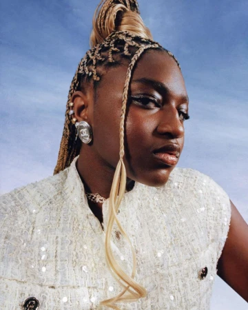 Zaya Wade wearing a textured off-white tweed top and silver logo earring against an open blue sky backdrop.