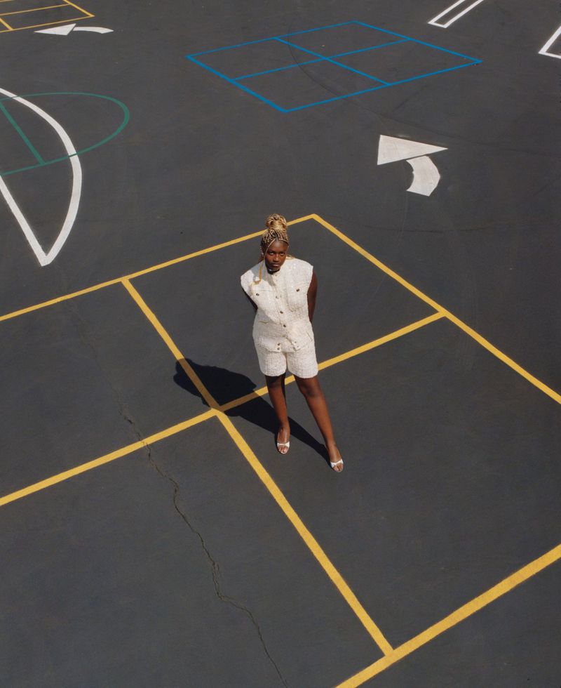 Overhead shot of a Black female standing on a paved surface with painted game markings wearing a cream-colored romper and heels.