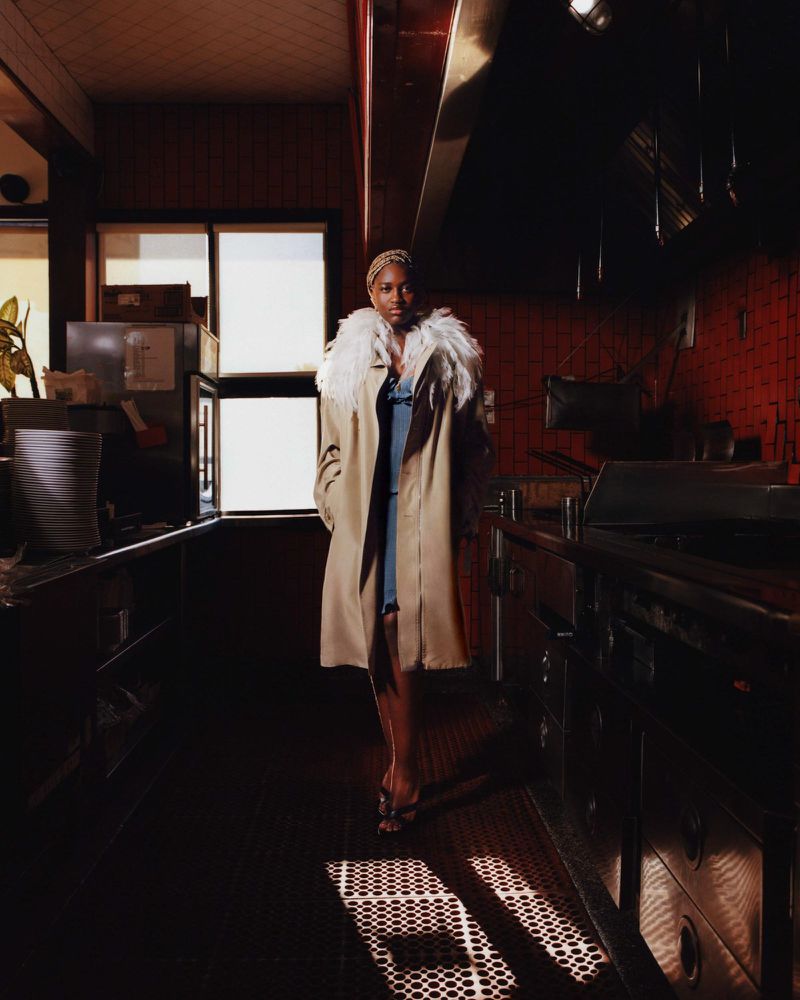 Woman in fur-collared coat walks through restaurant kitchen with dark tile floor and metal equipment under bright window light.