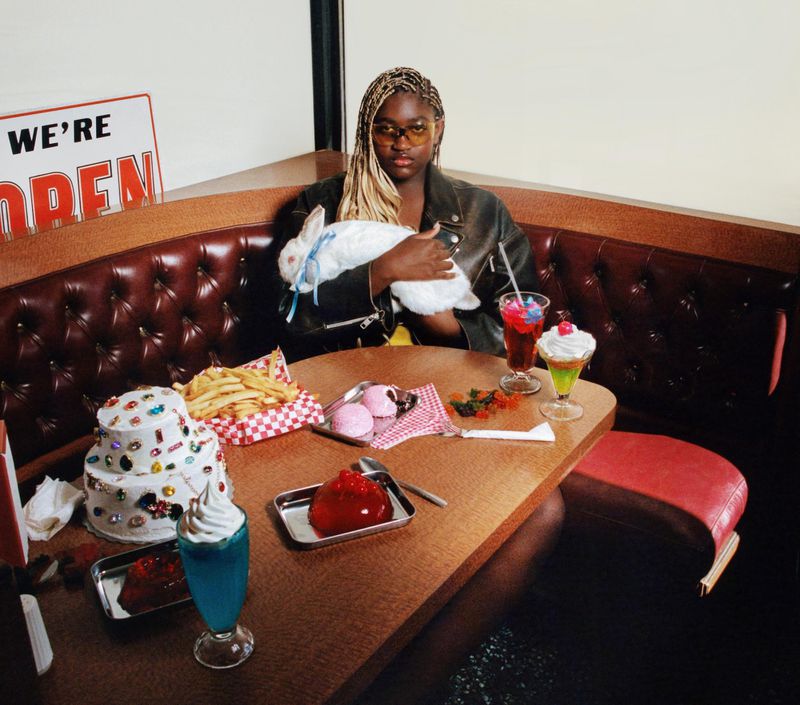 Young adult woman in booth with vintage diner desserts and pet rabbit, wears a leather jacket and retro sunglasses, posing indoors.