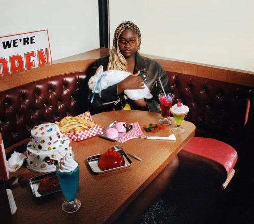 Young adult woman in booth with vintage diner desserts and pet rabbit, wears a leather jacket and retro sunglasses, posing indoors.