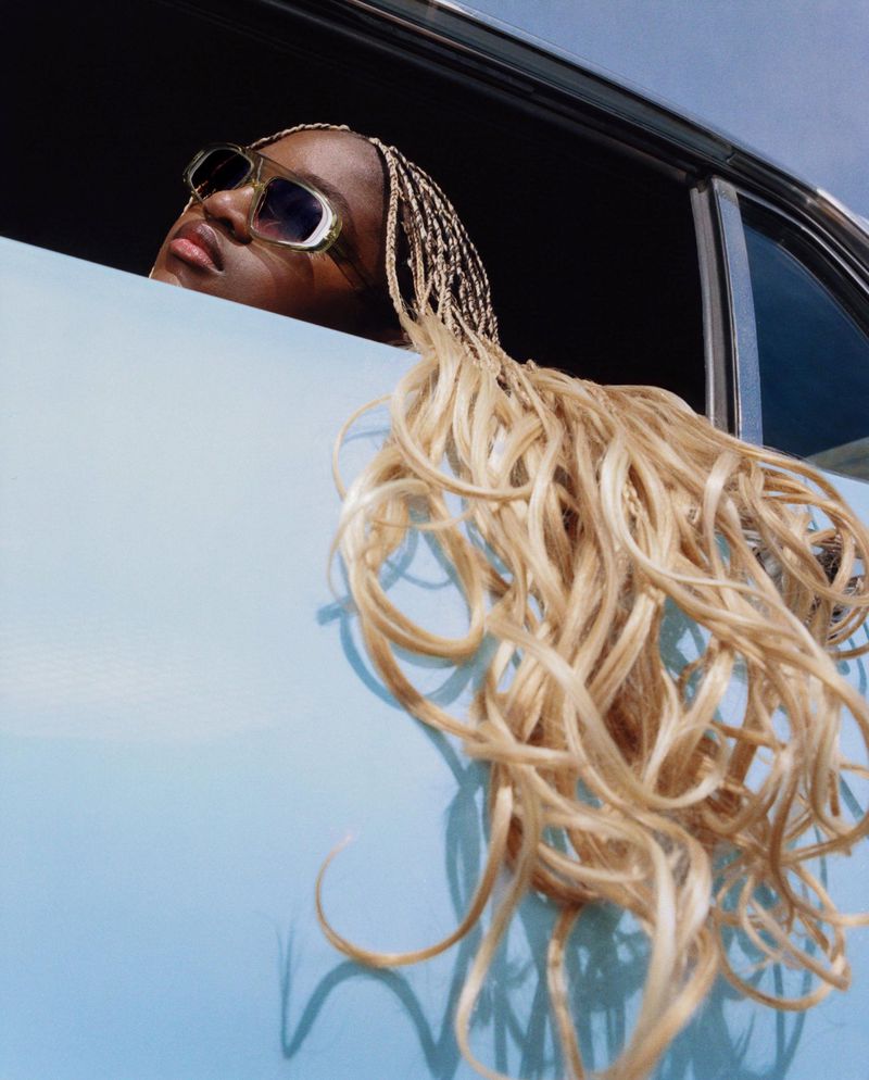 Stylish young woman with blonde braided hair leaning out of vintage car window, wearing sunglasses against a blue sky backdrop.