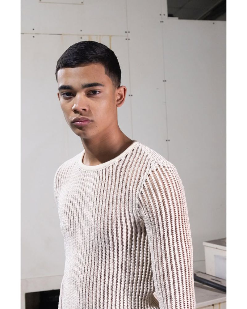 Portrait of a young adult male model wearing a beige knit sweater in a minimalist studio setting. Fashion editorial style.