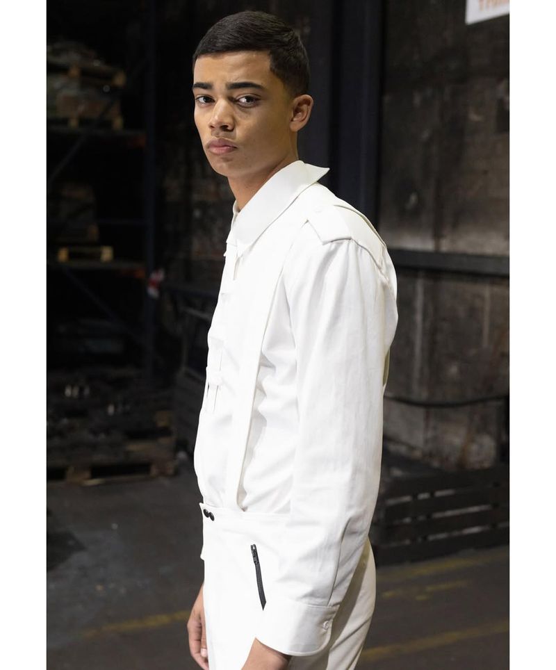 Young adult male in a white jacket and pants stands in a dimly lit industrial setting with metal shelves and neutral tones. 