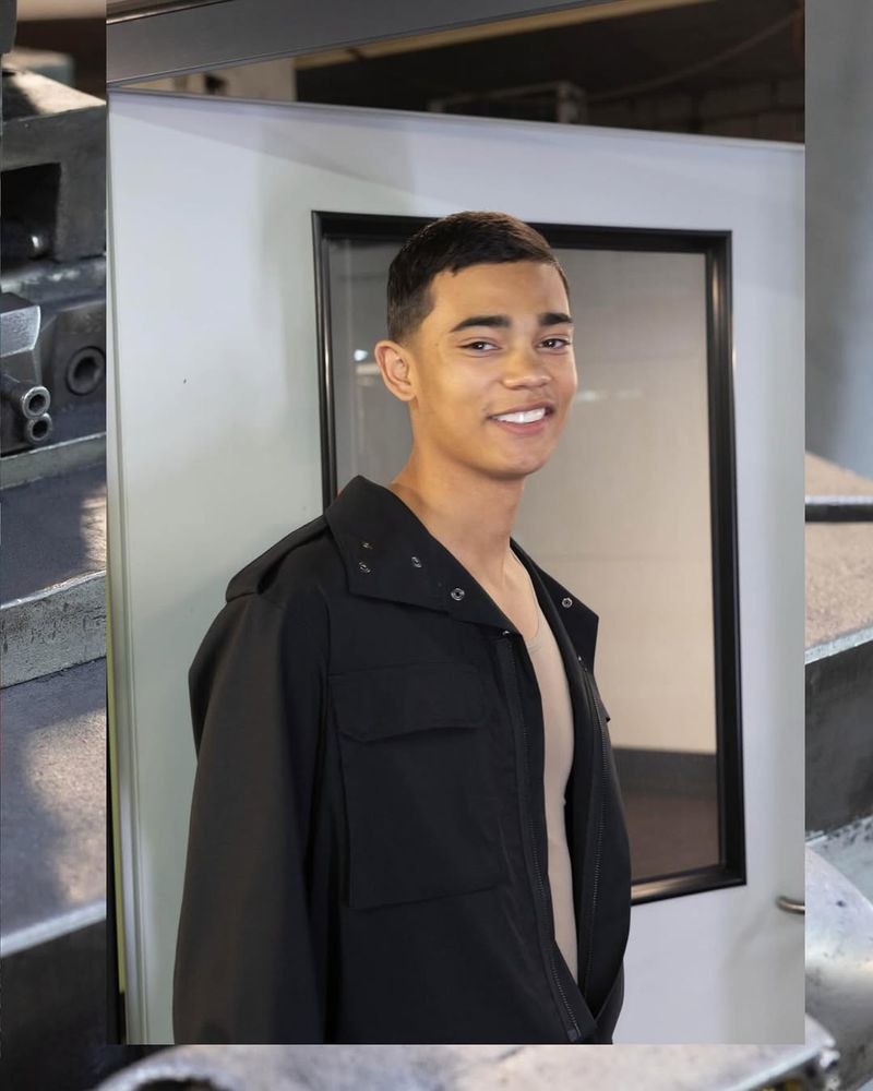 Smiling young man in a black jacket poses in front of a white door with metal machinery visible in the background in a studio setting.