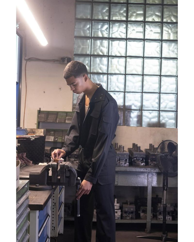Young adult using a vise in an indoor workshop, wearing a black jumpsuit, with a focus on tools and equipment, captured in a medium shot.
