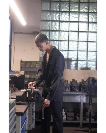 Model wearing dark utilitarian workwear standing beside a bench vise in an industrial workshop with glass block windows.