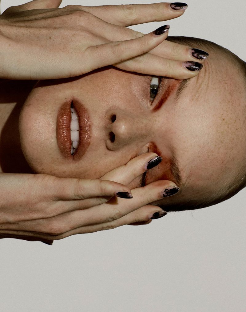 Close-up portrait of a woman's face partially obscured by her hands with dark nail polish against a white background.