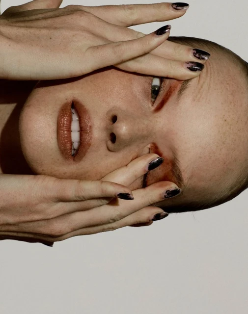 Close-up portrait of a woman's face partially obscured by her hands with dark nail polish against a white background. 