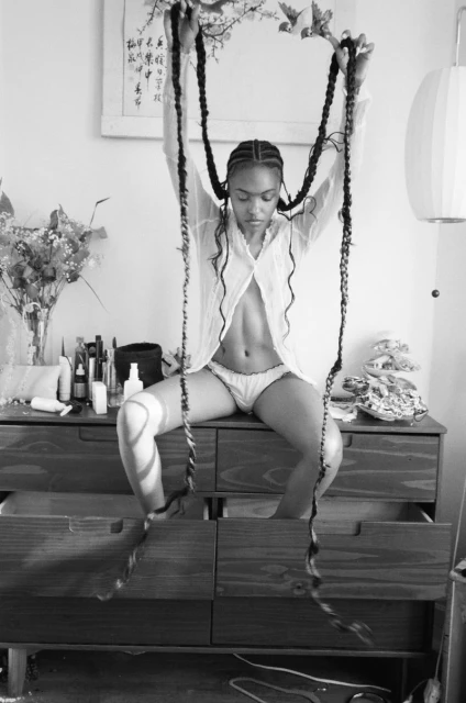 Black and white photo of a woman with long braids sitting on a wooden dresser, holding braids above her head, wearing white lingerie.