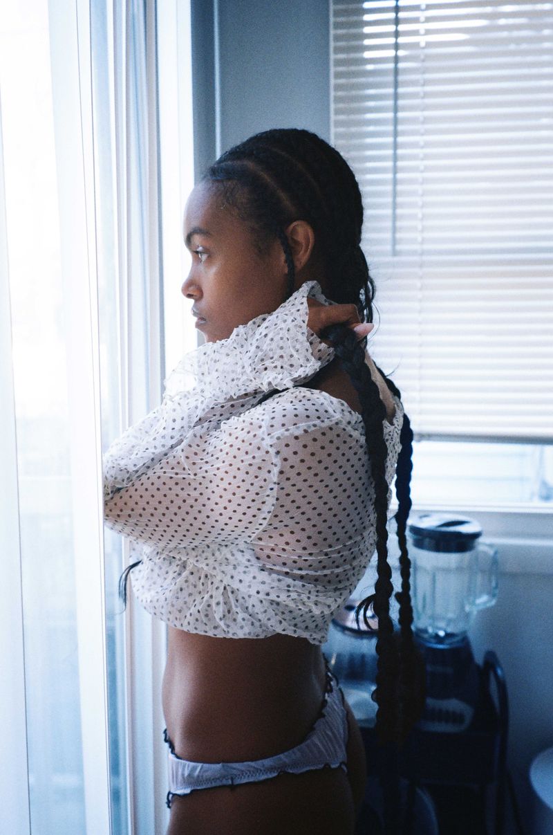 Young woman with braided hair in polka dot top stands indoors near a window with blinds, looking to the side in soft, diffused light.