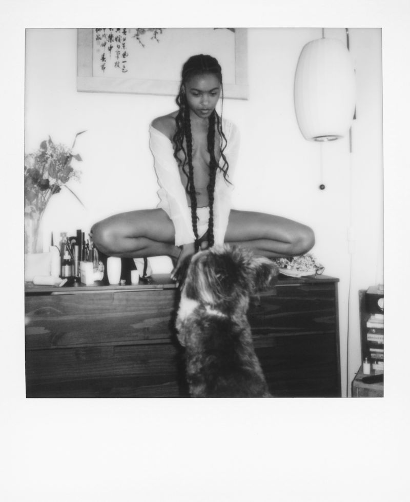 Monochrome portrait of a woman with long braids sitting on a dresser with various bottles and a dog looking up.