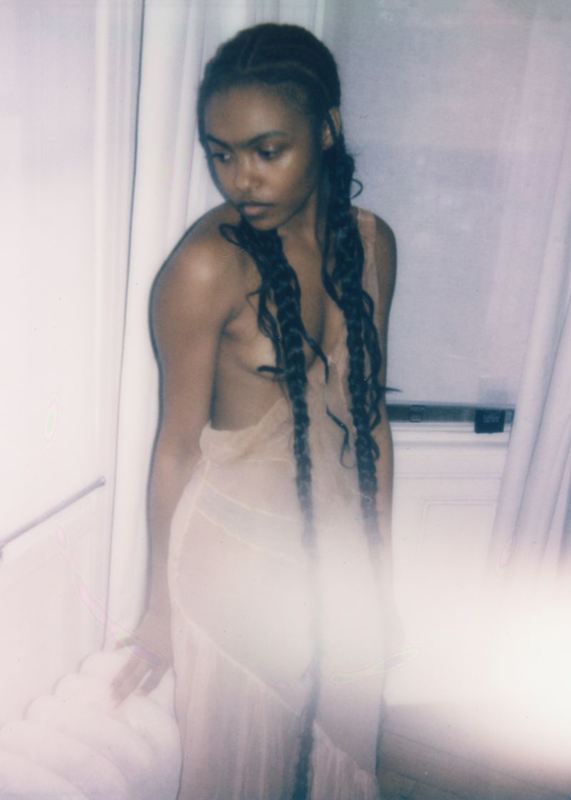 Portrait of an African-descent woman wearing a dress, posing indoors near a window with white curtains and diffused lighting.
