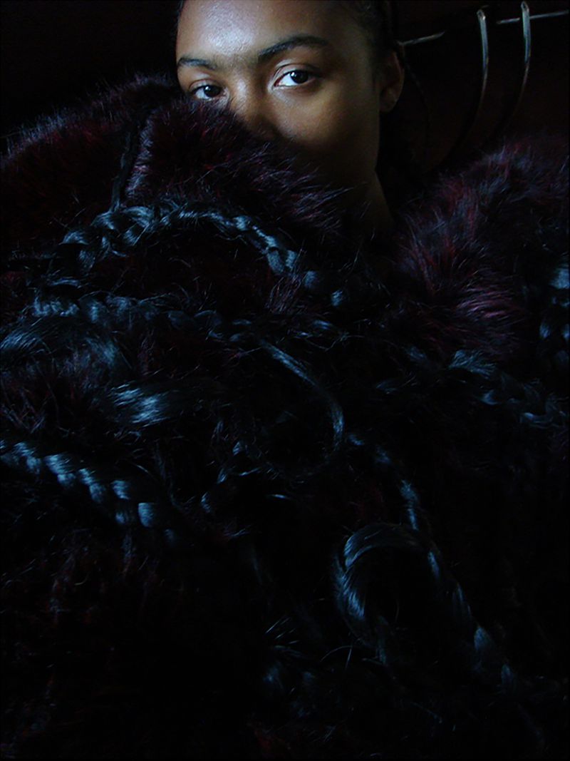 Close-up portrait of a woman enveloped in dark textured faux fur and braided hair details in low-key lighting.