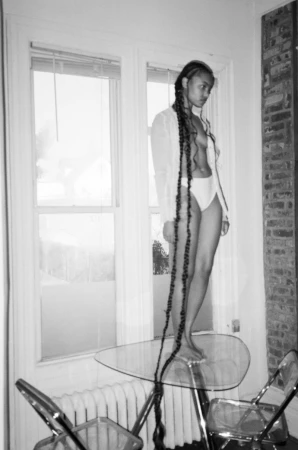 Adult female with long braids poses barefoot on a glass table in a white shirt and underwear with chairs and a window behind her.